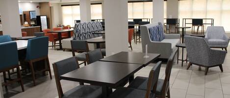 A spacious dining area with multiple tables and chairs, a counter with a coffee machine, and large windows providing natural light.