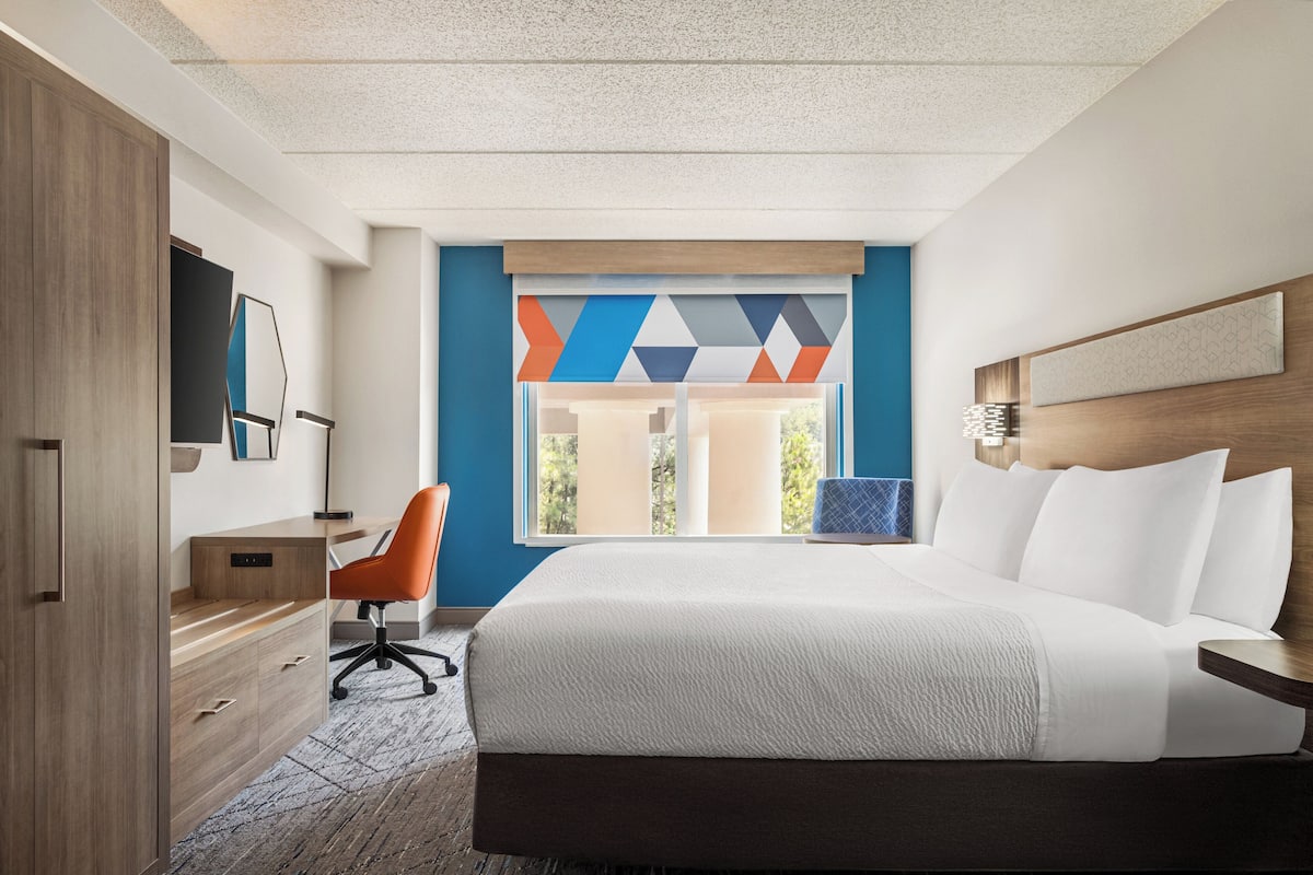 A hotel room with a bed, a desk, a chair, a TV, and a window with a geometric view.