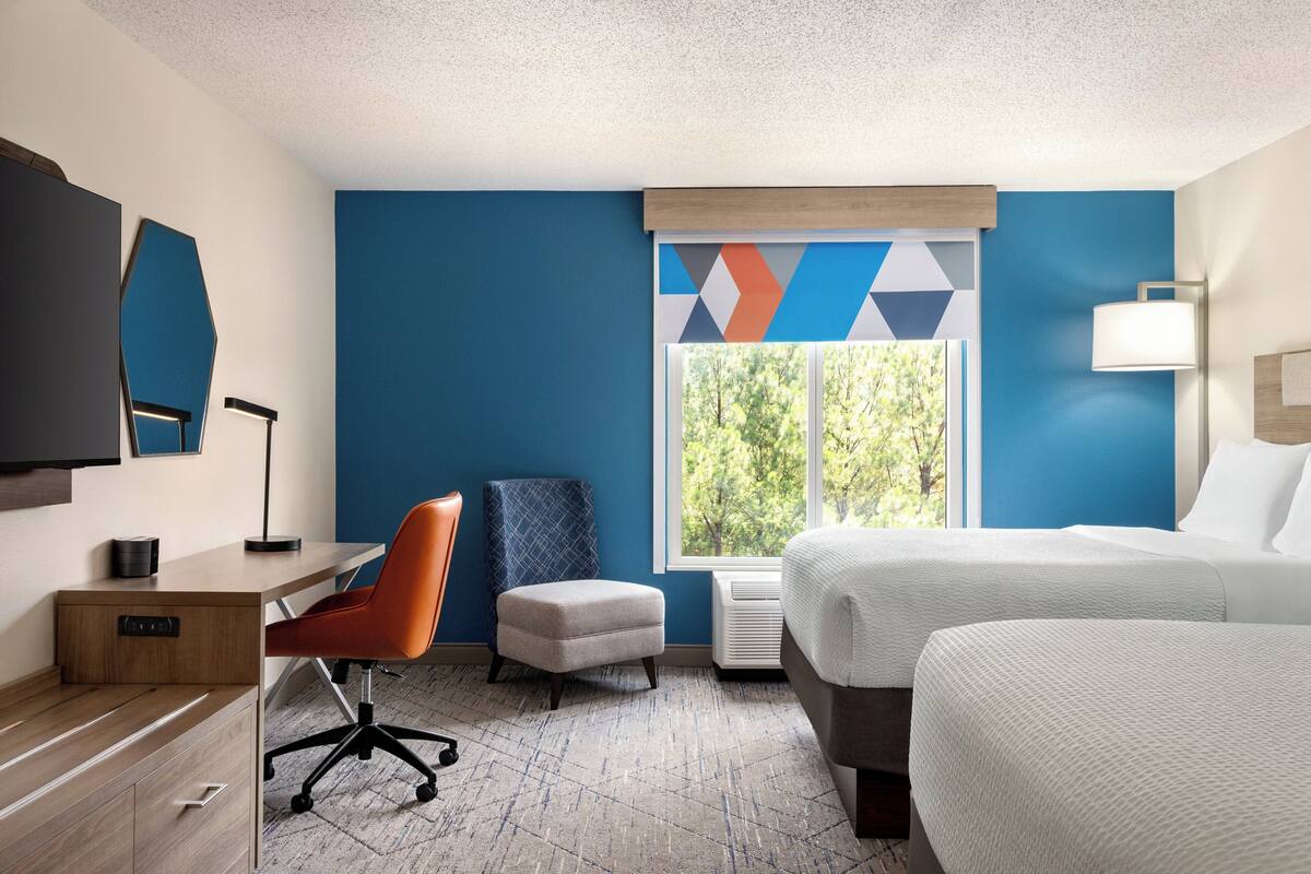 A hotel room with a bed, desk, chair, and a large window with a view of trees.