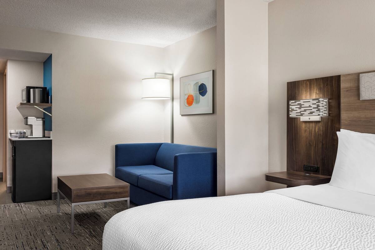 A hotel room with a bed, a blue sofa, a small table, a desk with a computer, and a wall-mounted lamp.
