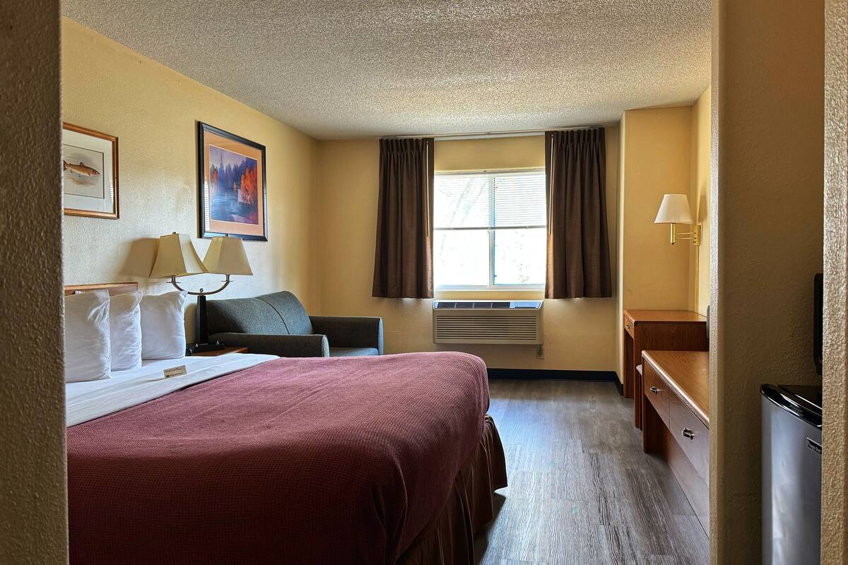 A hotel room with a bed, a desk, a chair, a window with curtains, a lamp, and a wall-mounted air conditioner.