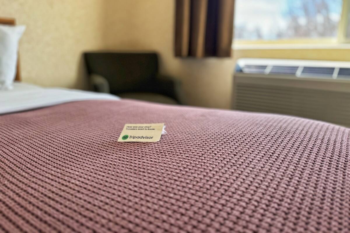 A hotel room with a bed, a chair, a window with curtains, and a wall-mounted air conditioning unit.