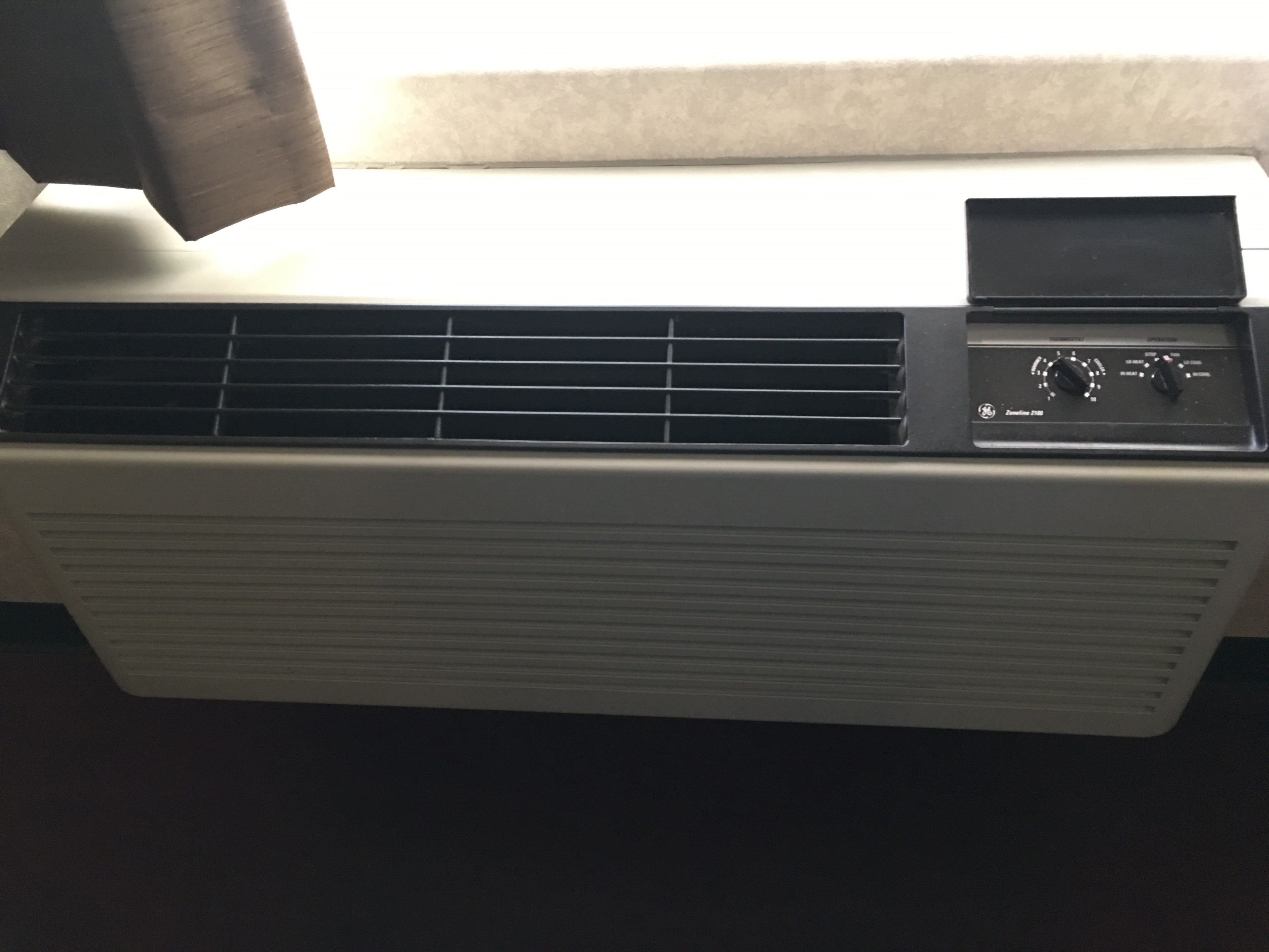 A white portable air conditioner with a control panel, placed near a window with a white blind.