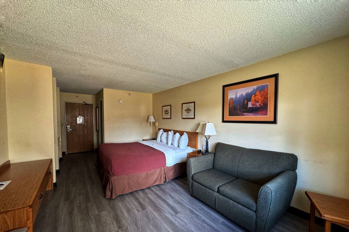 A hotel room with a bed, a gray sofa, a wooden desk, and a framed picture on the wall.