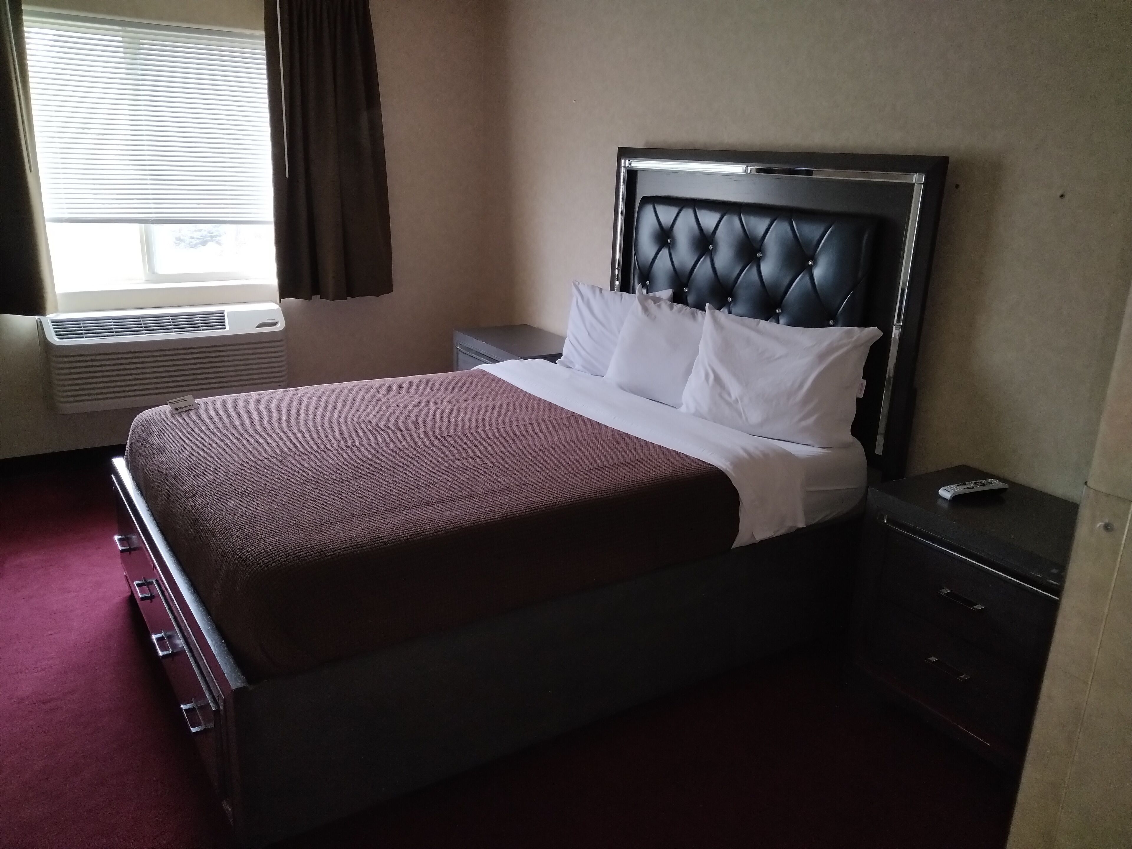 A neatly made bed with a dark headboard, a bedside table with a remote control, and a window with blinds.