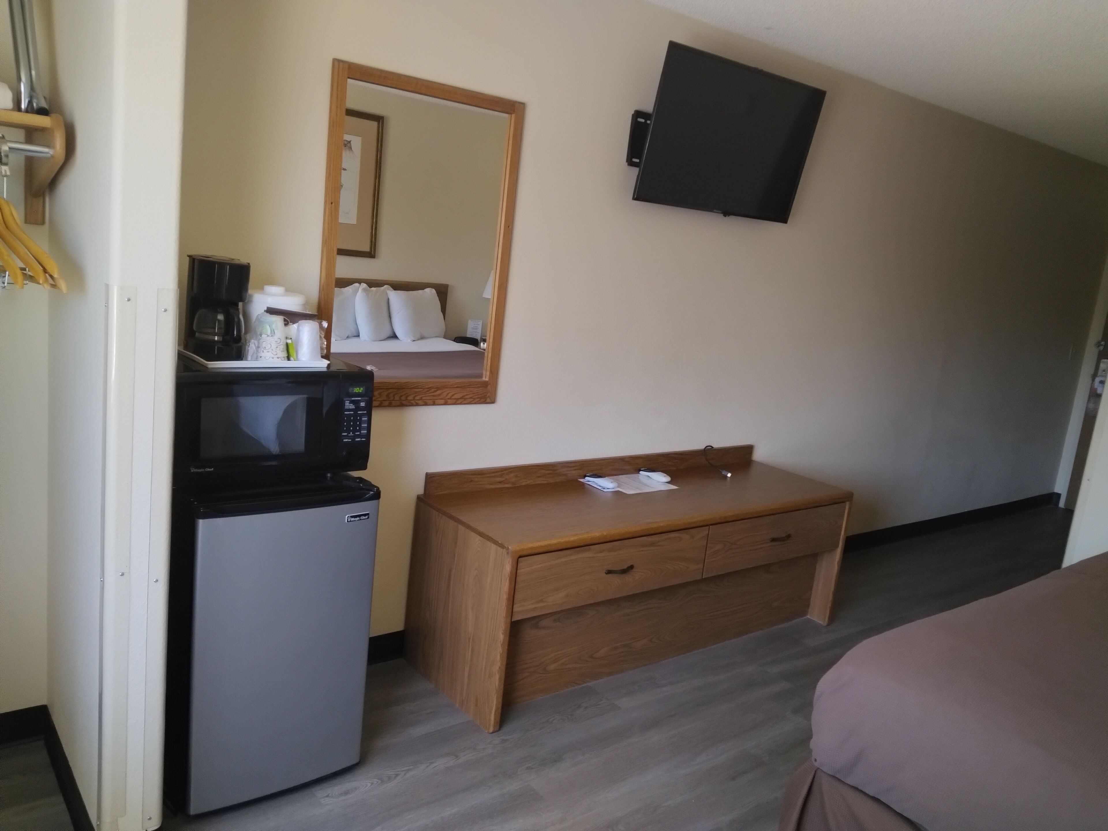 A compact hotel room with a kitchenette area including a microwave, refrigerator, and coffee maker. A wooden dresser and a flat-screen TV are also visible.