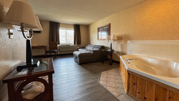 A hotel room with a wooden floor, a sofa, a small table with a lamp, a bathroom with a bathtub, and a window with curtains. - Stay Wise Inn Cedaredge (Cedaredge)