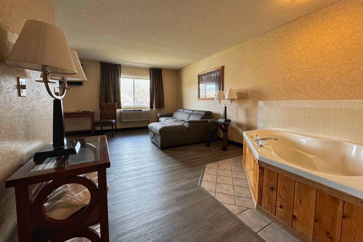 A hotel room with a wooden floor, a sofa, a small table with a lamp, a bathroom with a bathtub, and a window with curtains.