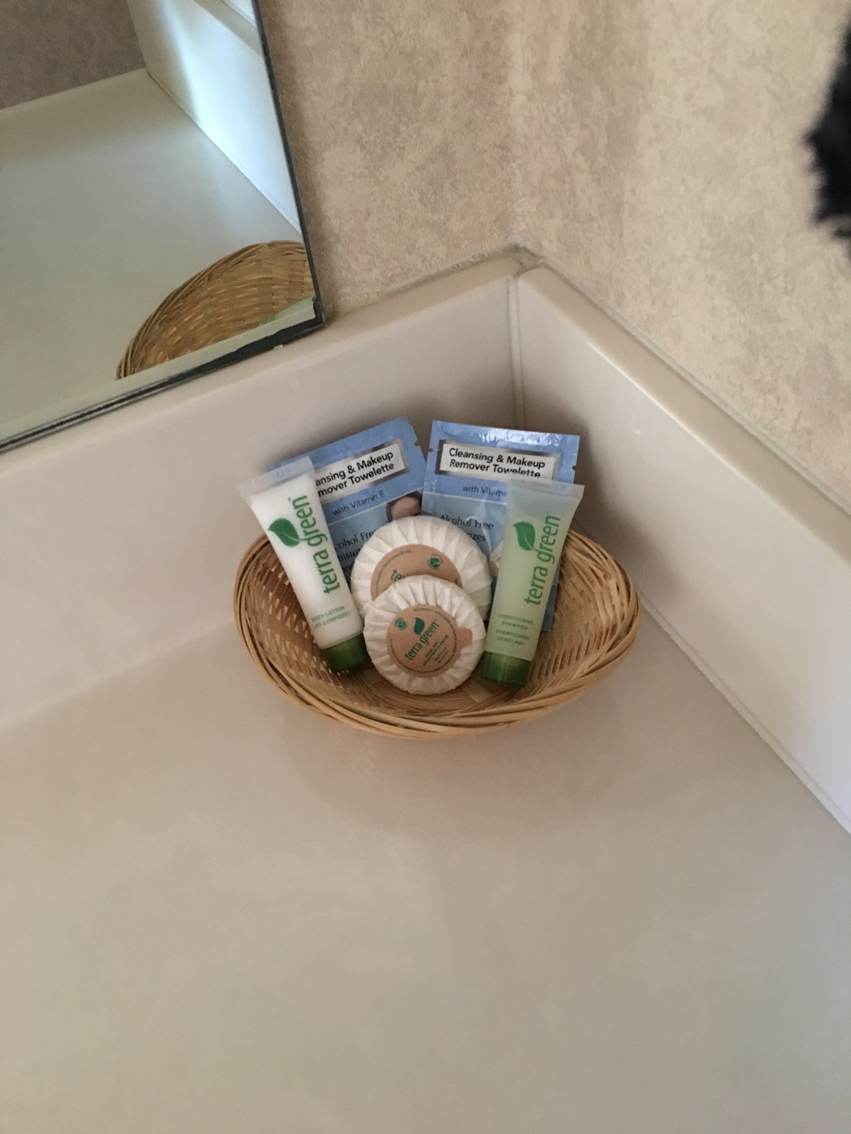 Een badkamermuis met een rieten mand, gevuld met toiletartikelen zoals een reinigingsmelk, make-up remover en shampoo.