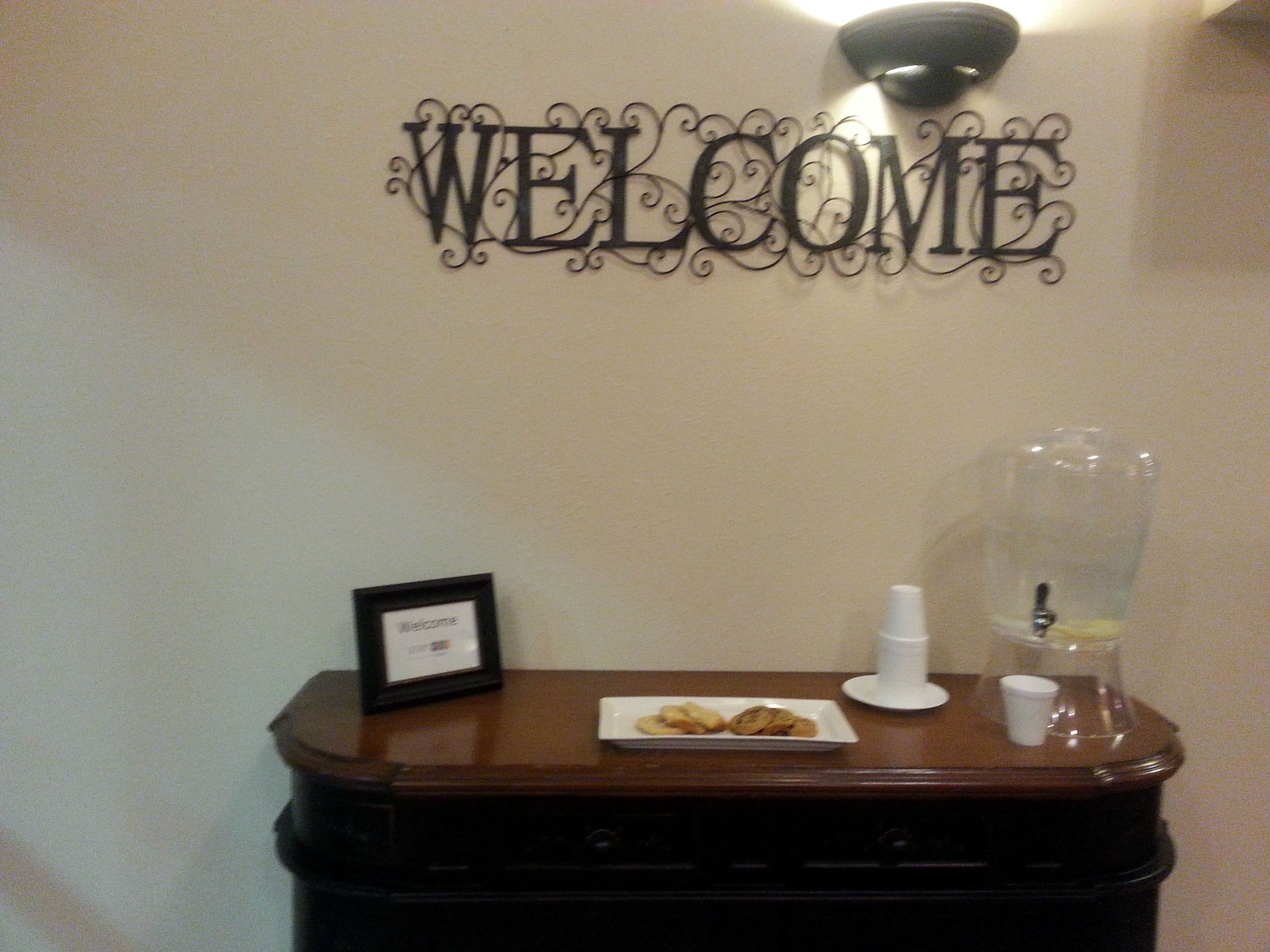 A welcome sign, a framed welcome card, a plate with cookies, a glass pitcher, and a cup on a wooden table.