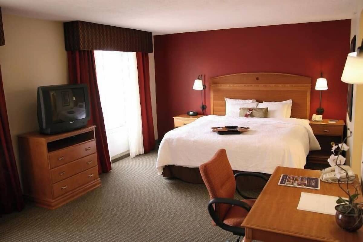 Une chambre d’hôtel avec un lit, un téléviseur posé sur une commode en bois, un bureau avec une chaise et une table de chevet avec des lampes.