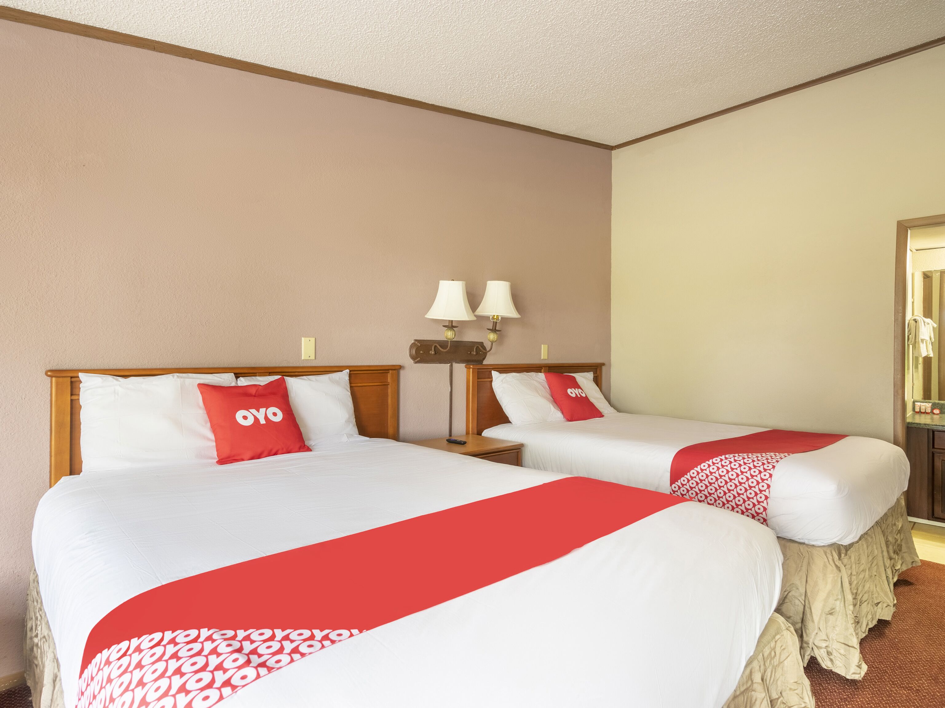 A hotel room with two beds, each with white linens and red accents, a nightstand with lamps, and a mirror.