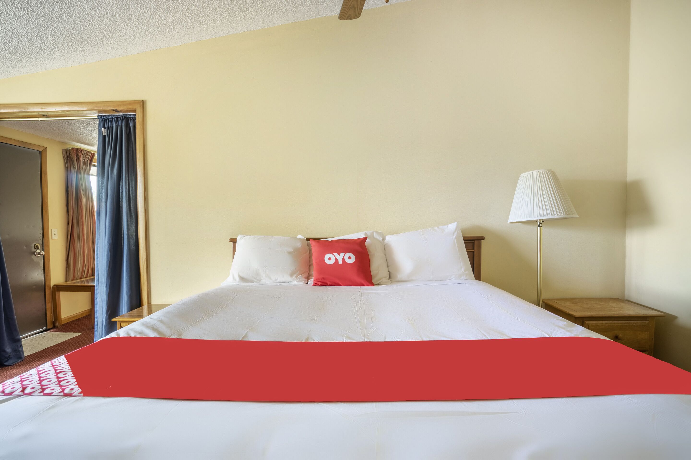 A hotel room with a bed, a nightstand, a lamp, and a door with curtains.