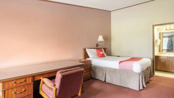 A hotel room with a single bed, a desk with drawers, a chair, a nightstand with a lamp, and a bathroom with a mirror and sink.
