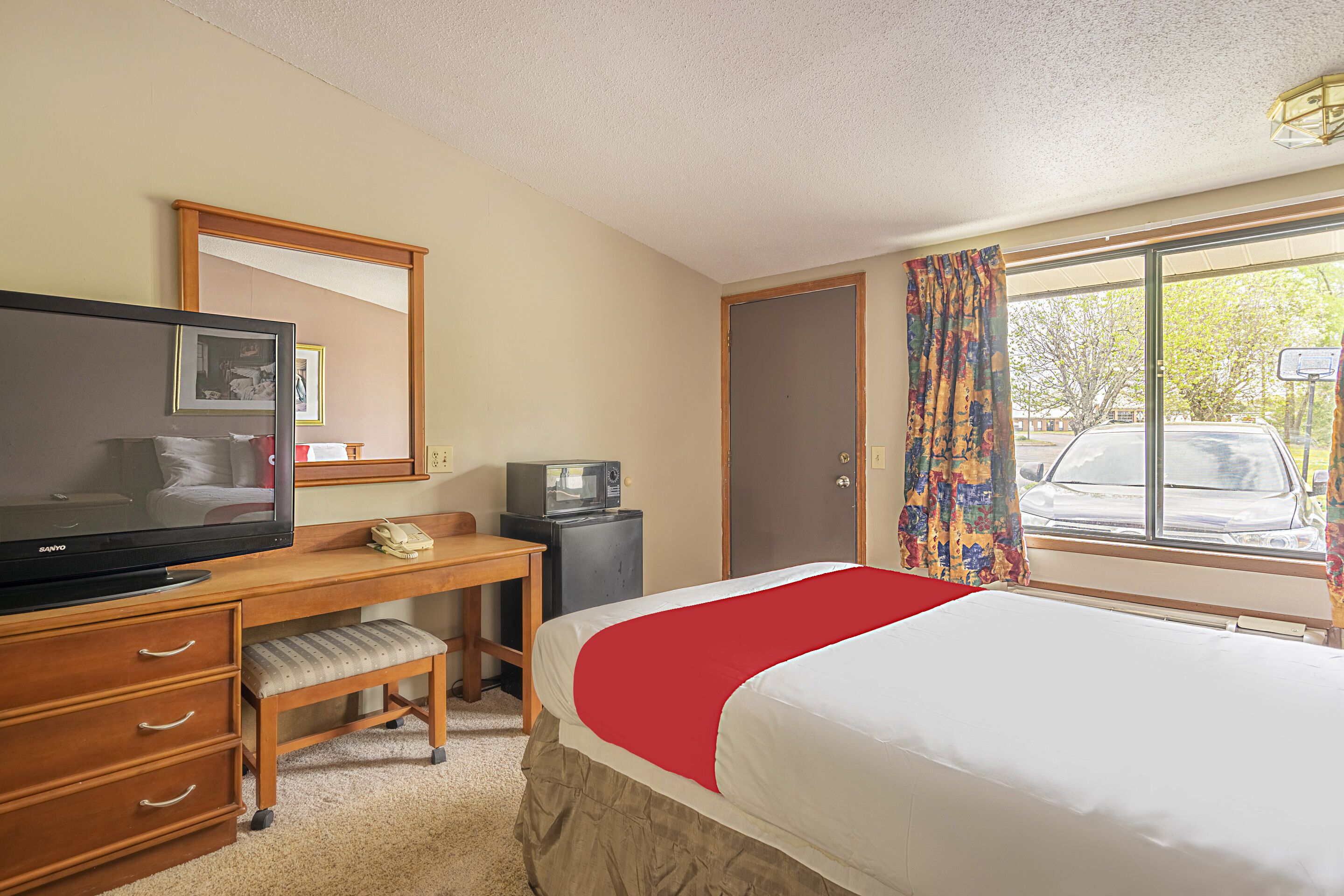 A hotel room with a bed, a desk with a television, a telephone, and a small refrigerator.
