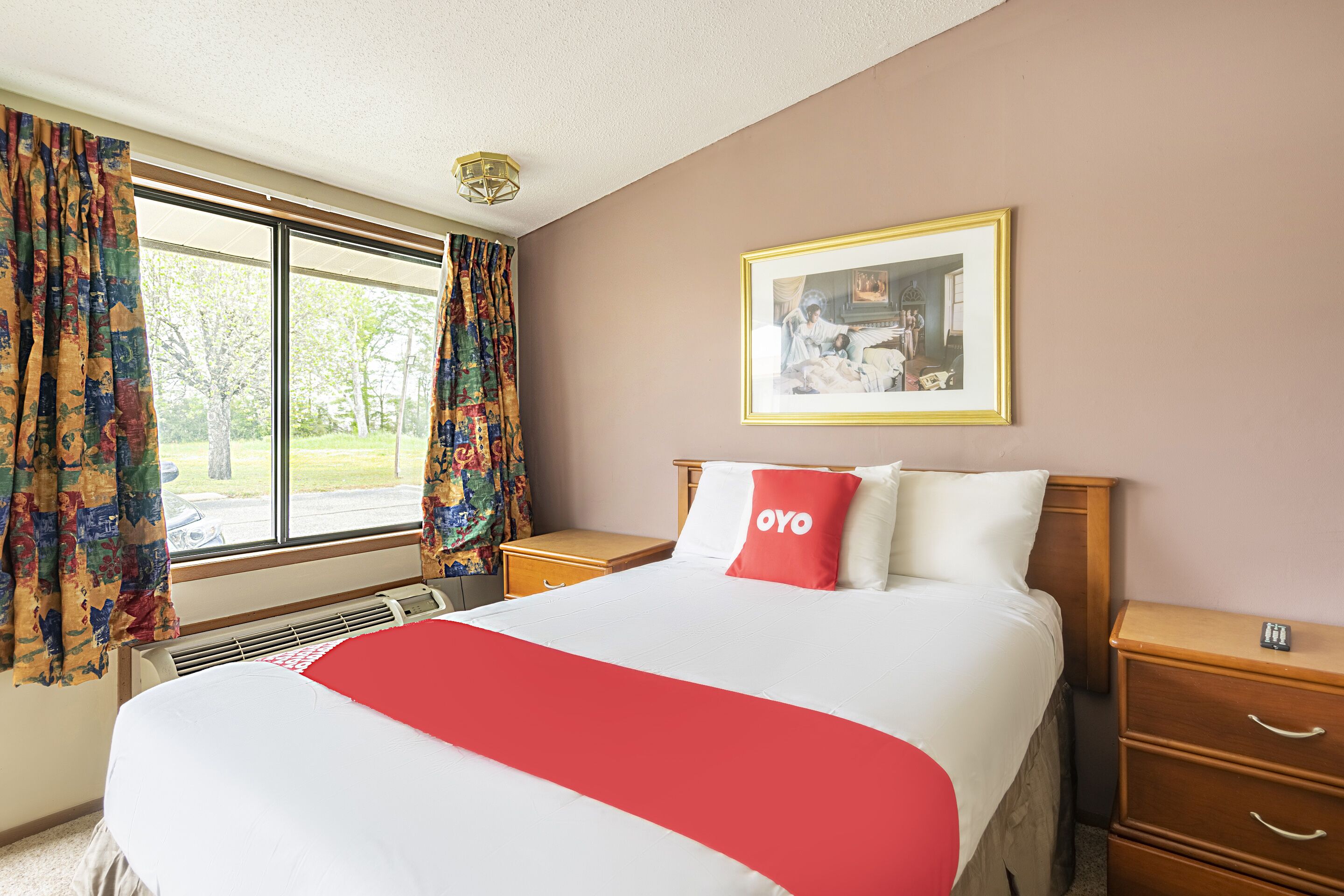 A hotel room with a large bed, a bedside table, a nightstand, a window with curtains, a wall-mounted picture, and a ceiling light.