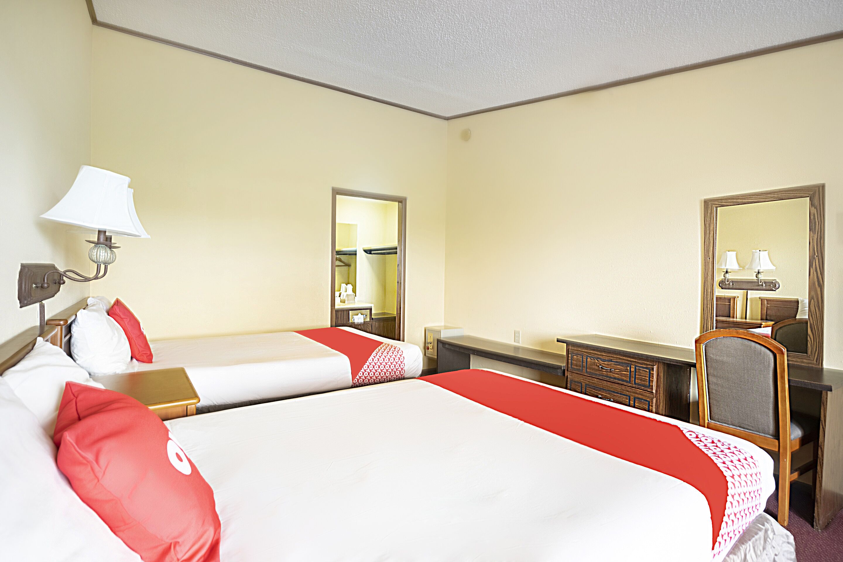 A hotel room with two beds, a desk, a chair, a mirror, and a lamp.