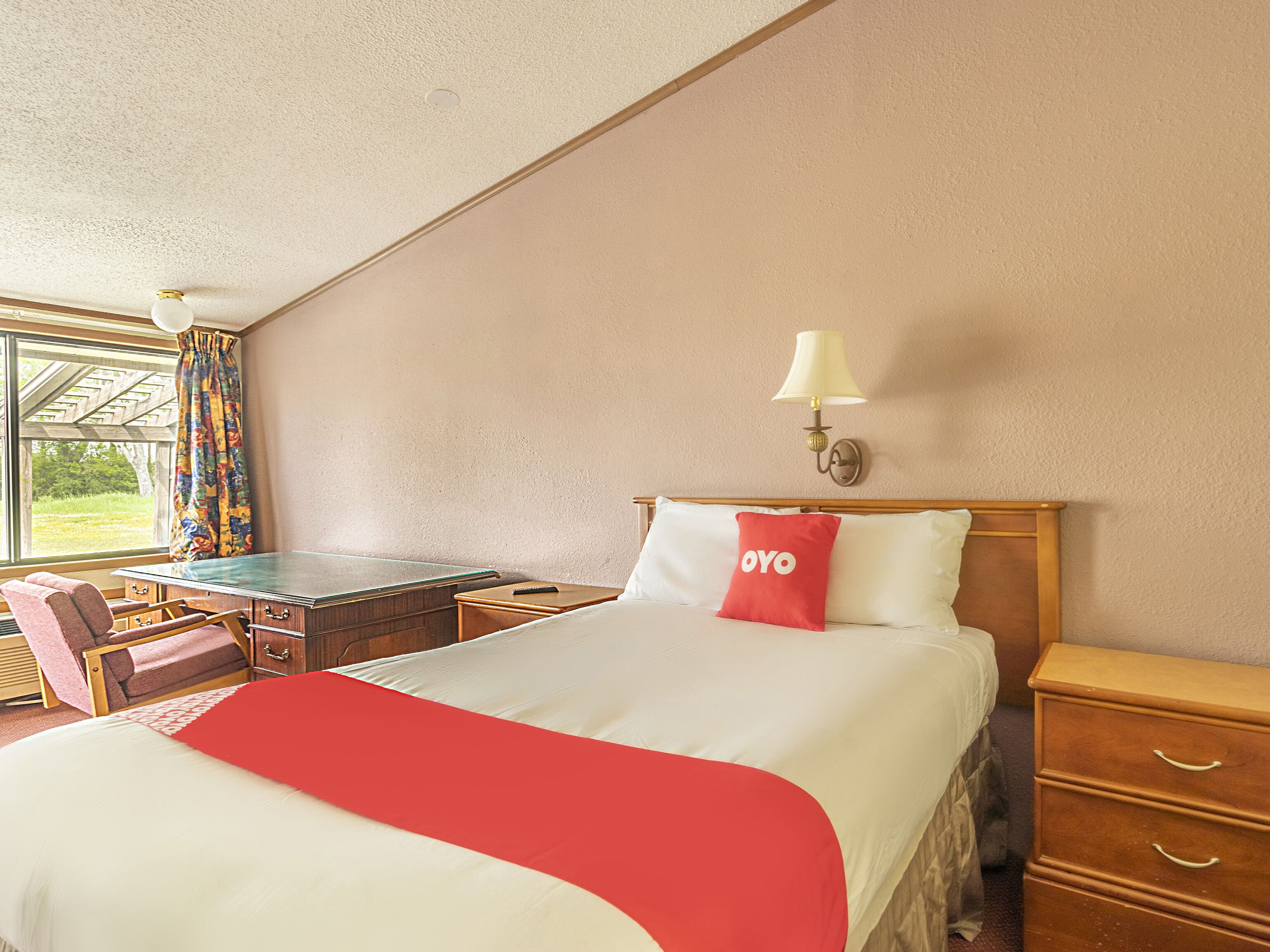 A hotel room with a bed, a desk, a chair, a lamp, and a window with curtains.