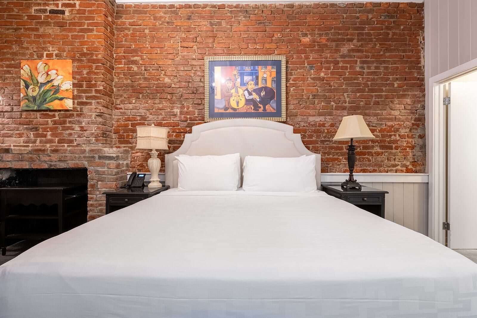 Habitación de hotel con pared de ladrillo, una cama grande con ropa blanca, un cuadro sobre la cama y una chimenea a la izquierda.