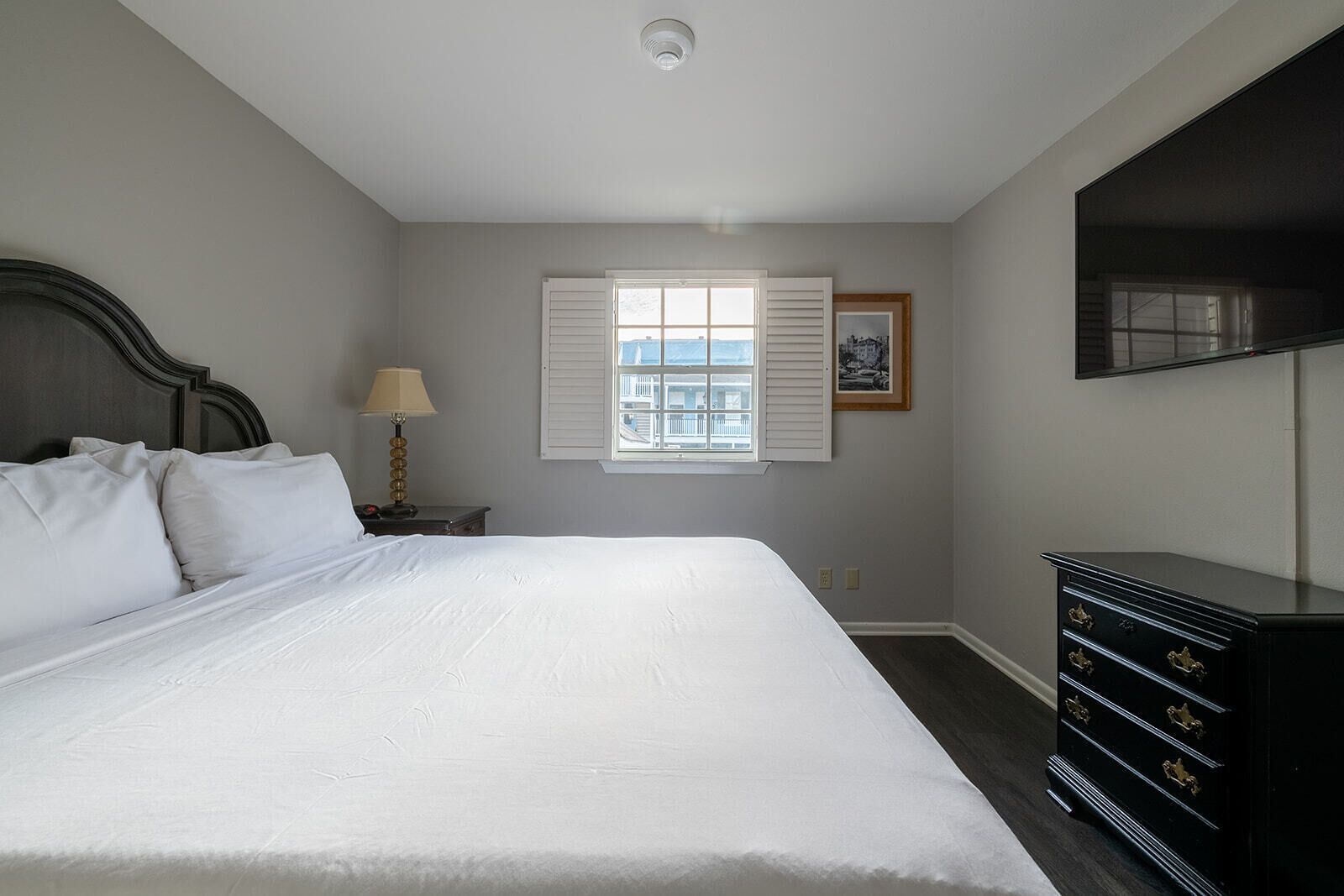 Une chambre avec un grand lit, une table de chevet avec une lampe, une commode, une fenêtre avec des volets et un tableau encadré au mur.
