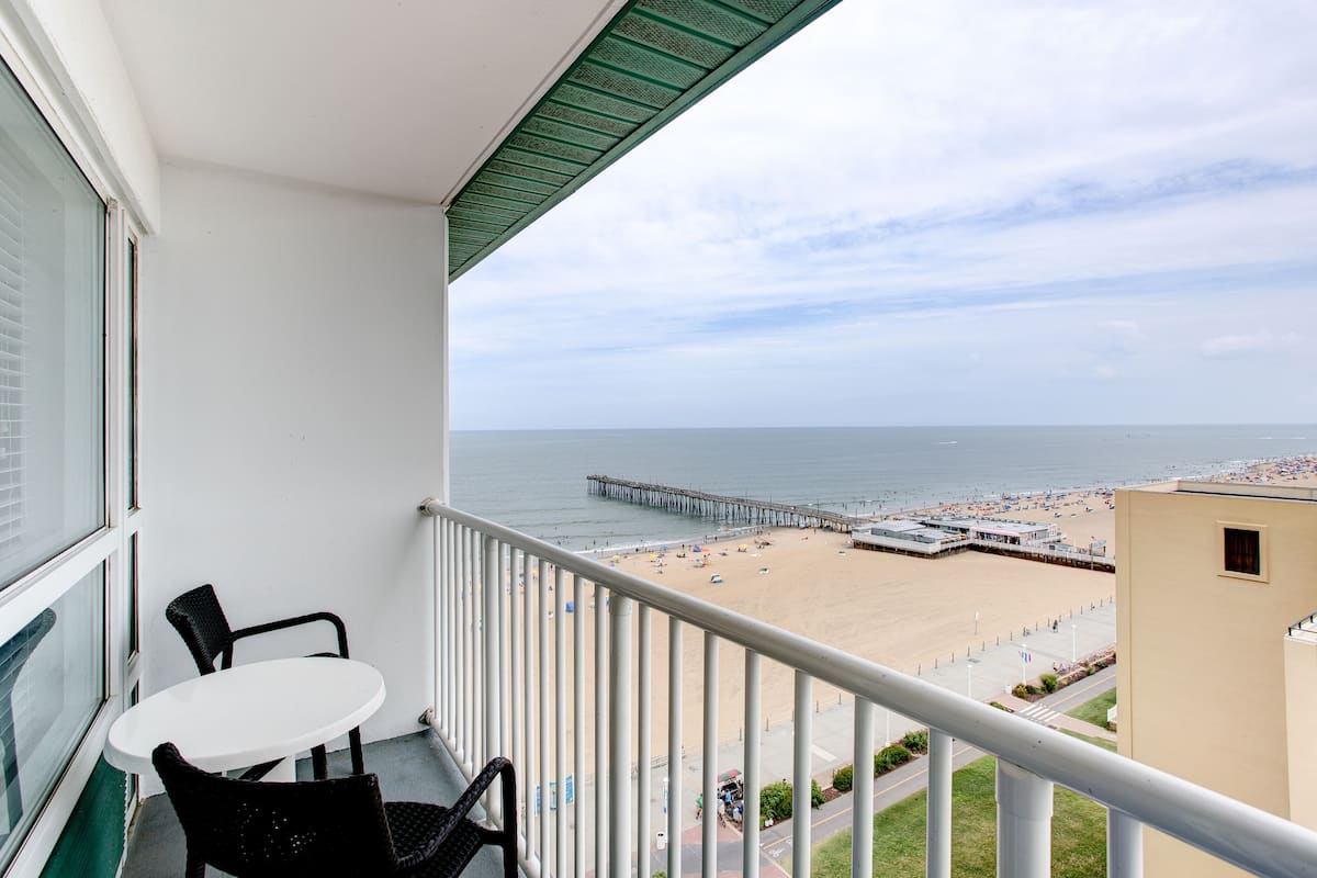 A balcony with a white table and two chairs, overlooking a beach with a pier and a boardwalk.