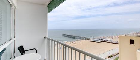 A balcony with a white table and two chairs, overlooking a beach with a pier and a boardwalk.