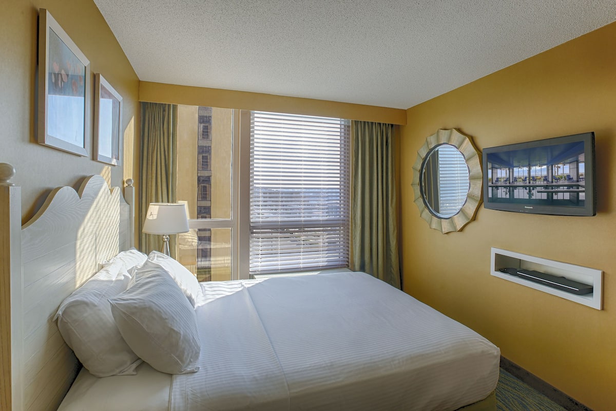 A hotel room with a large bed, a television, a mirror, and a window with blinds.