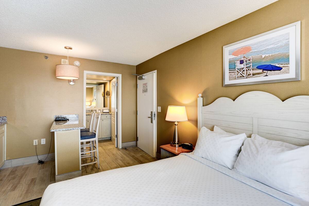 A hotel room with a bed, a desk with a chair, a nightstand with a lamp, and a painting of a beach scene.