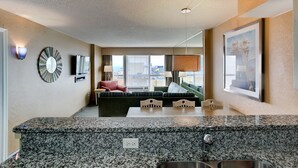 A modern hotel room with a kitchen area, a seating area with a sofa and chairs, and a large window with a city view. - Boardwalk Resort and Villas (Virginia Beach)