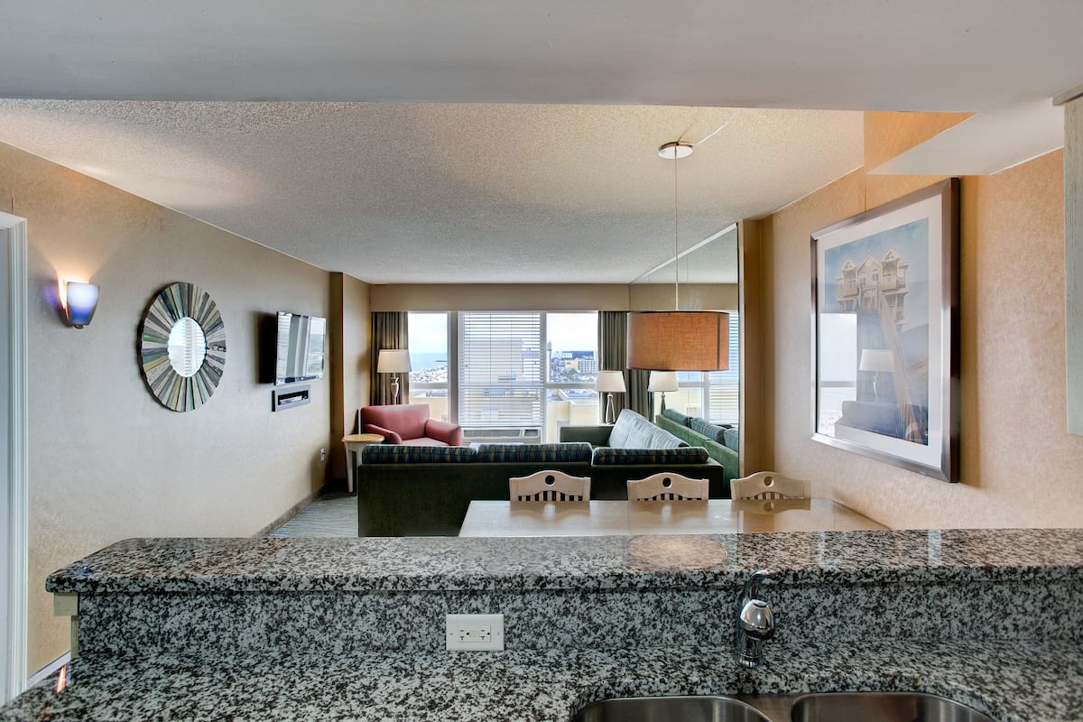A modern hotel room with a kitchen area, a seating area with a sofa and chairs, and a large window with a city view.