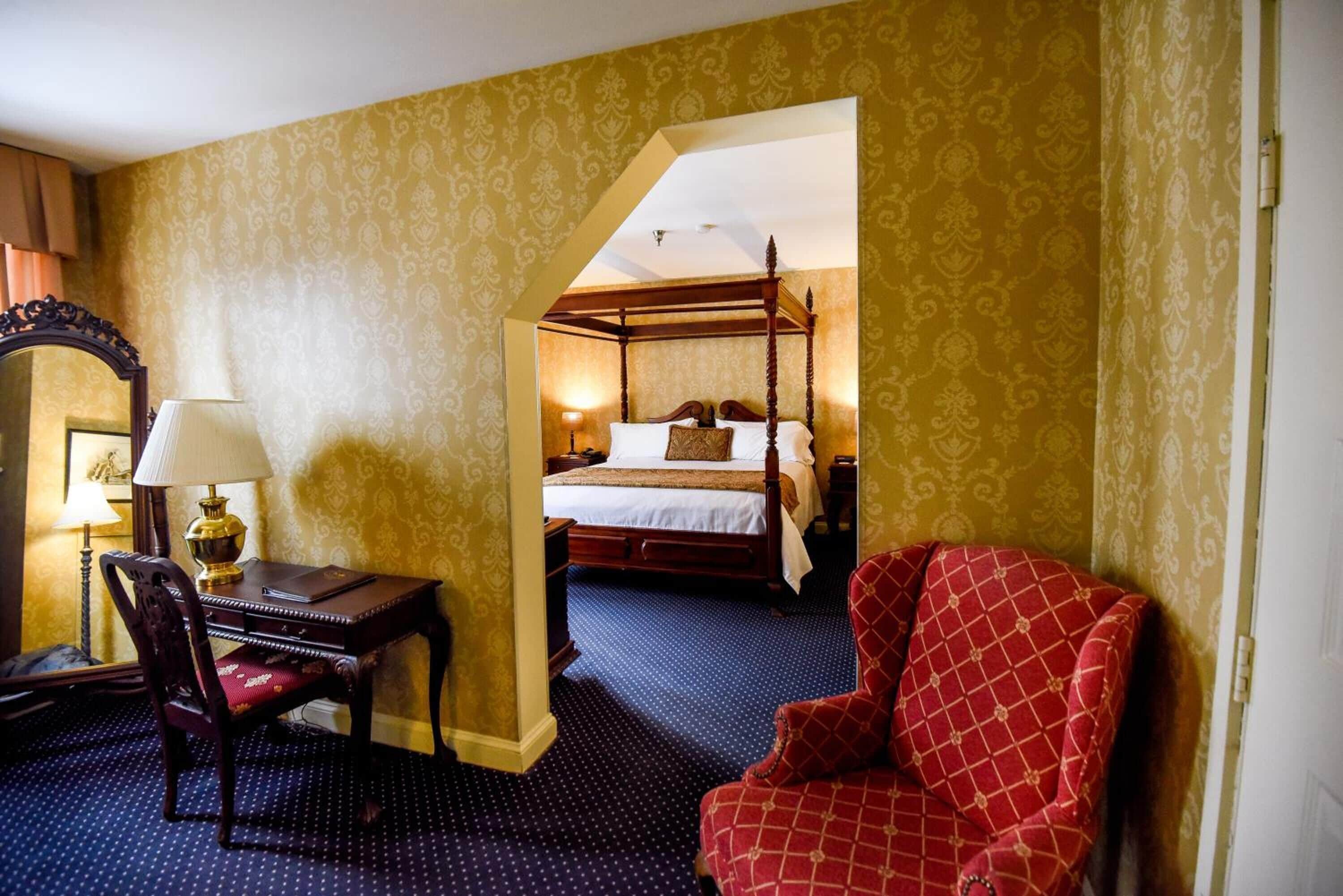 Una habitación de hotel con una cama con dosel, un escritorio con lámpara y un sillón rojo.