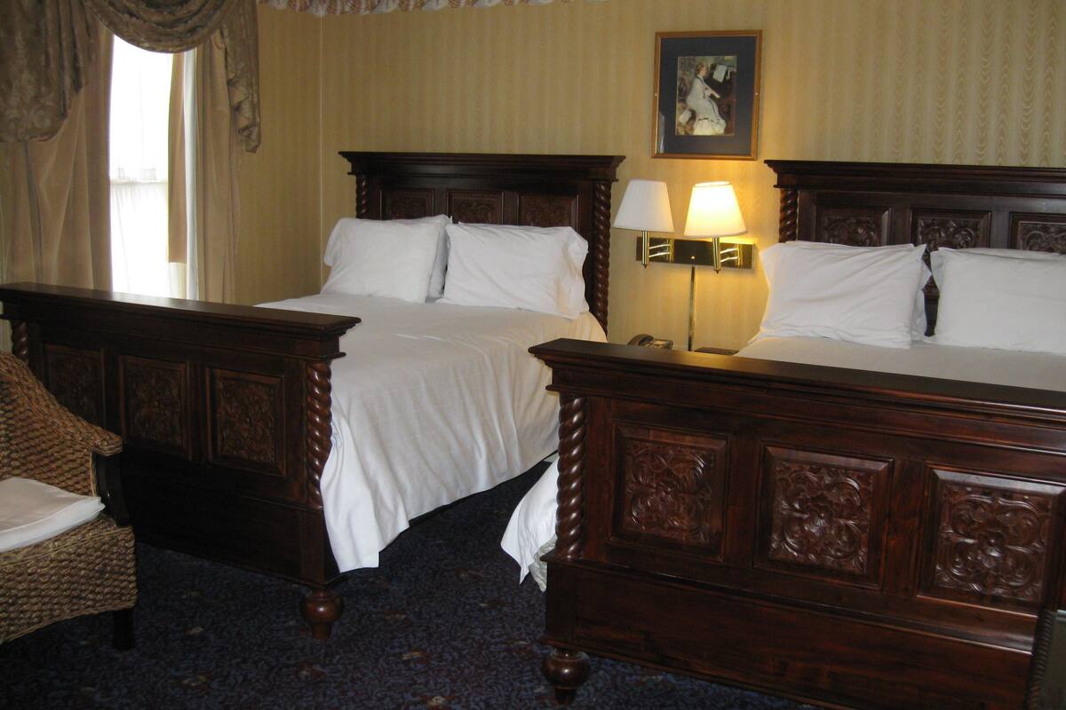 A hotel room with two beds, a wooden headboard, a nightstand with a lamp, a chair, and a framed picture on the wall.