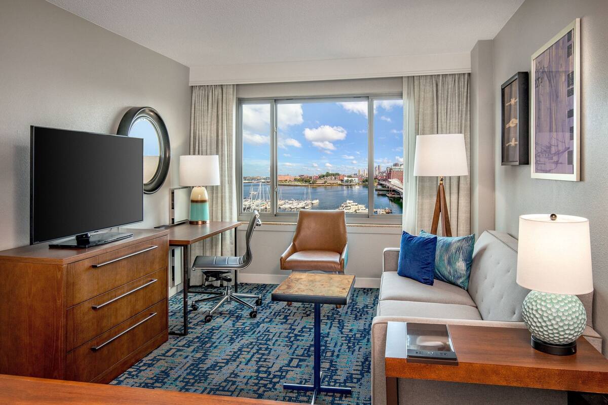A hotel room with a large TV, a desk with a lamp, a sofa with a lamp, a chair, and a view of a harbor.