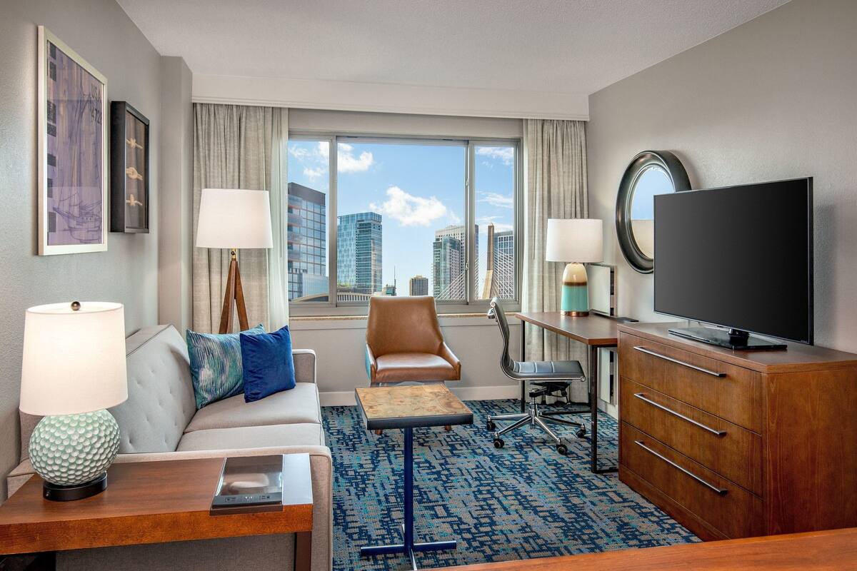 A hotel room with a sofa, armchair, desk, television, and a large window offering a city view.