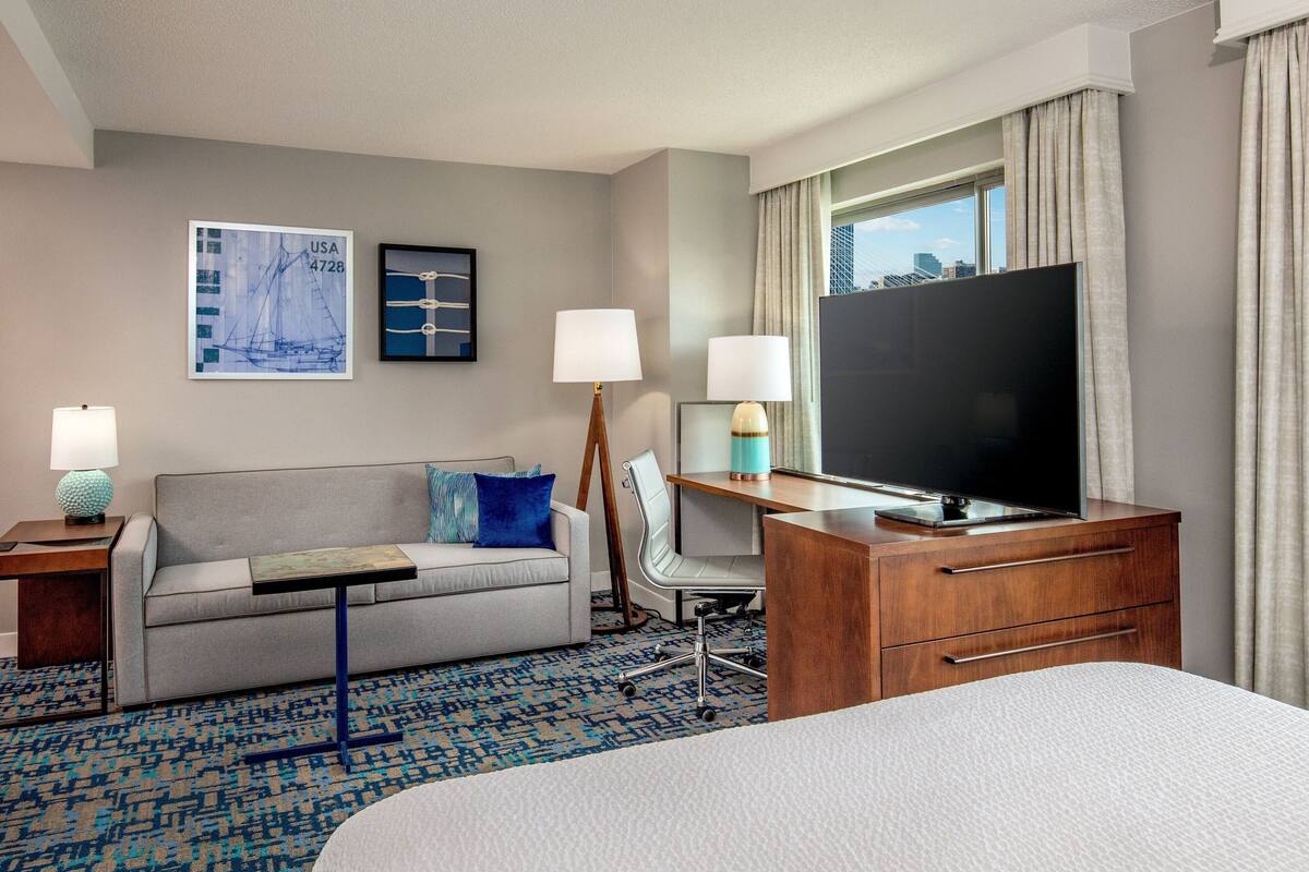 A hotel room with a sofa, a desk with a TV, a lamp, a chair, and a large bed.