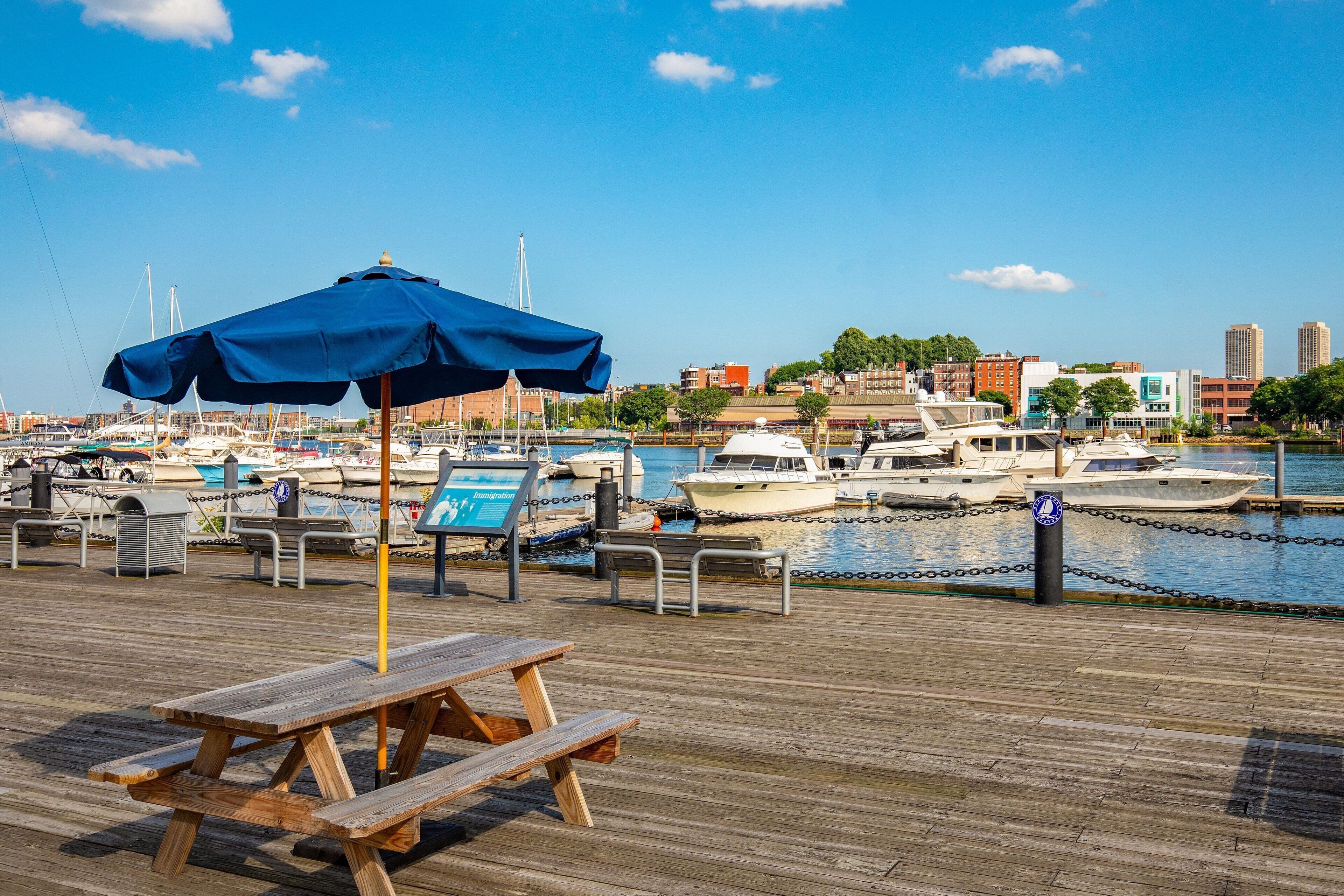 Un muelle de madera con mesas de picnic y una sombrilla azul, con vistas a un puerto deportivo con barcos y edificios al fondo.