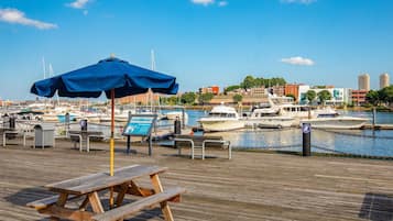 Un muelle de madera con mesas de picnic y una sombrilla azul, con vistas a un puerto deportivo con barcos y edificios al fondo.