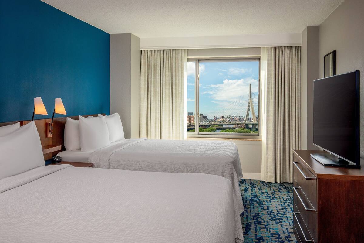 A hotel room with two beds, a TV, a desk, and a large window with a city view.