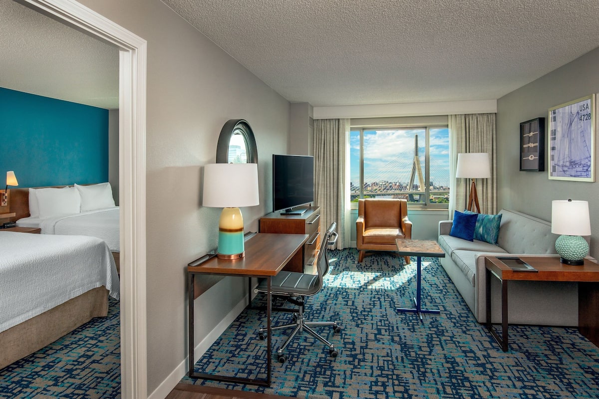 A hotel room with a bed, a desk with a lamp, a chair, a sofa, a TV, and a large window with a view of a bridge.