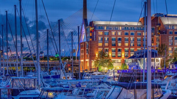Un port de plaisance où sont amarrés de nombreux bateaux, un bâtiment moderne à la façade en briques et, au loin, un pont à haubans.