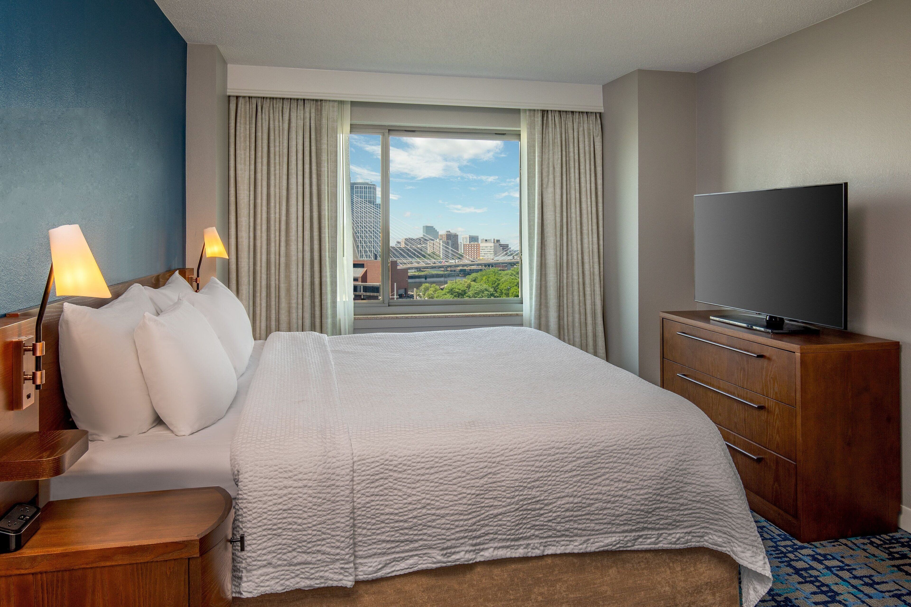 A hotel room with a large bed, a TV on a wooden cabinet, and a view of a cityscape through a large window.