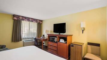 A hotel room with a bed, a desk with a chair, a television, a microwave, a cabinet, and a lamp.