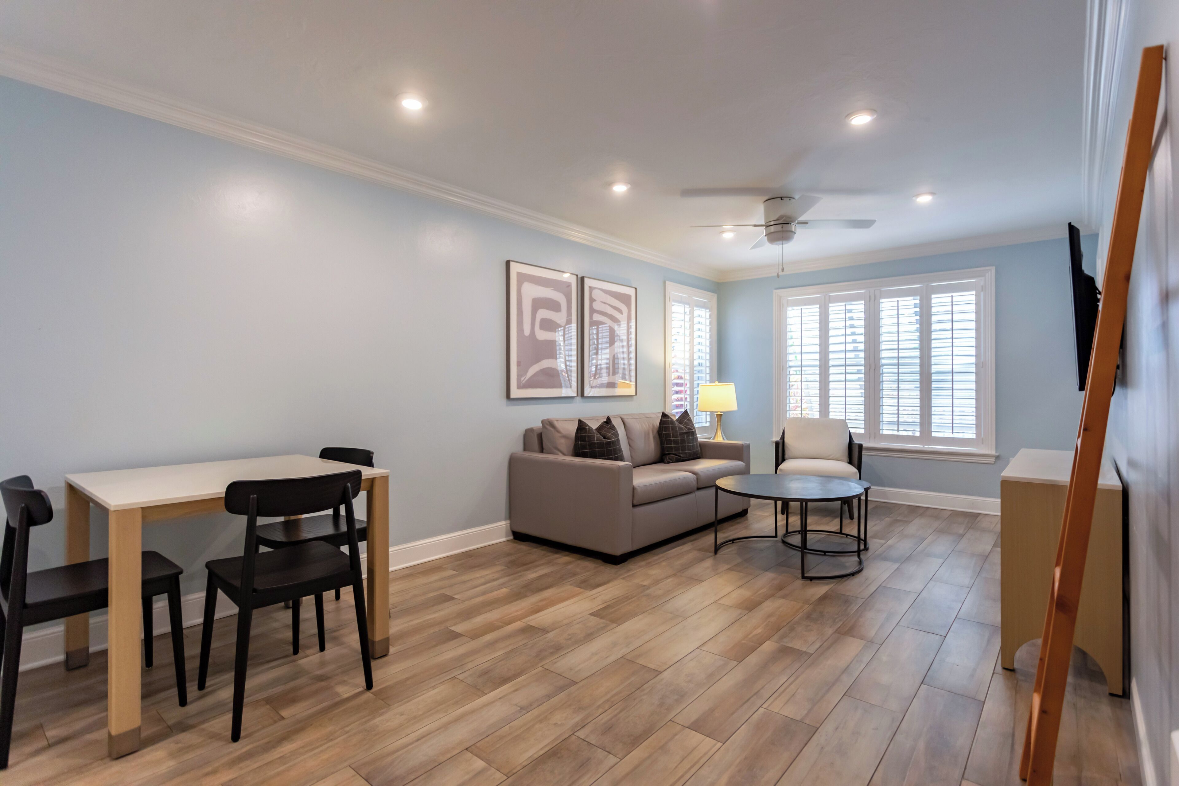 Un salon avec un canapé beige, une table basse et une table à manger avec des chaises. Il y a deux tableaux encadrés au mur, un ventilateur de plafond et une fenêtre avec des stores.
