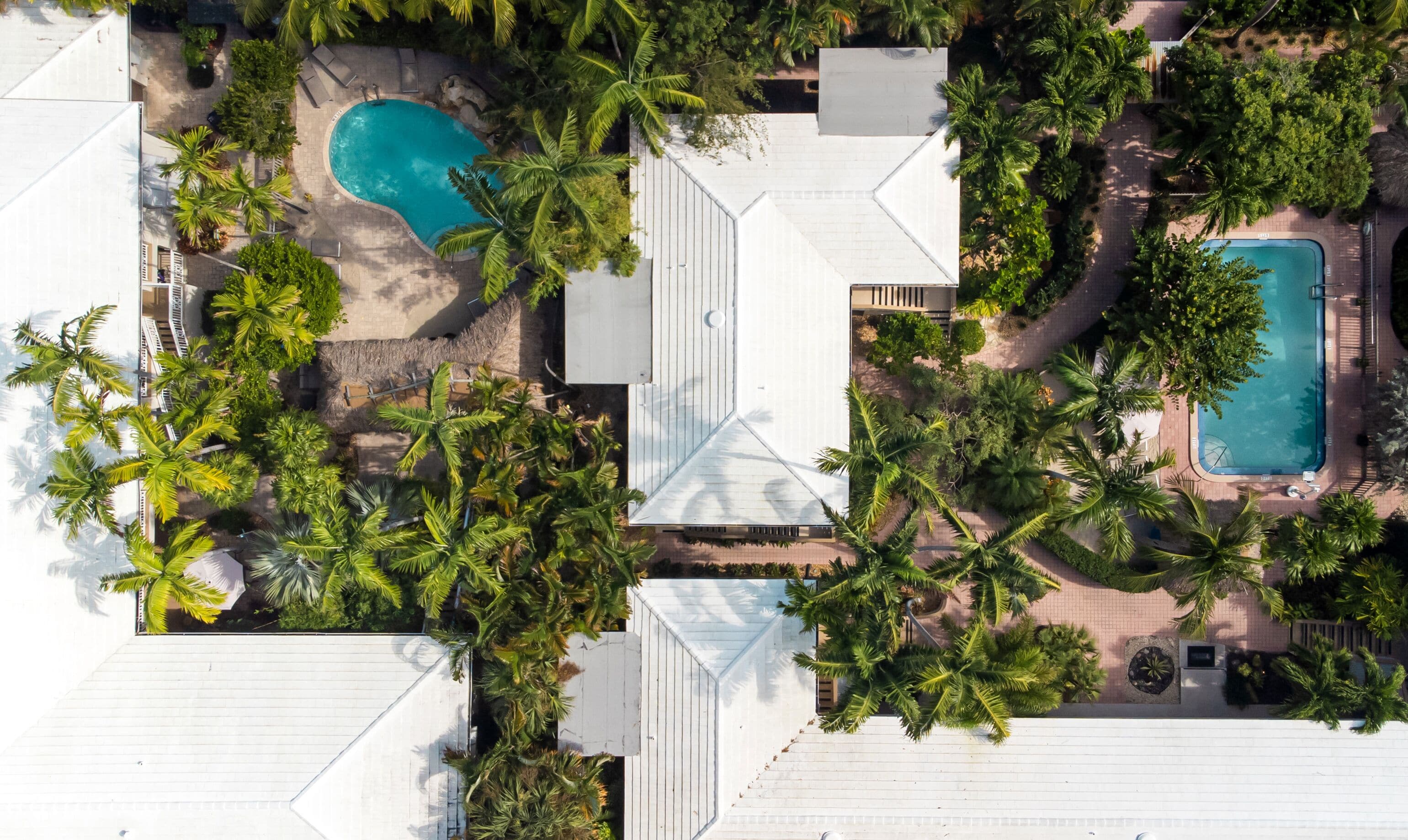 Vue aérienne d’un complexe hôtelier doté de deux piscines, entouré de palmiers et d’un bâtiment à toit blanc.