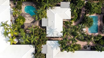 Vue aérienne d’un complexe hôtelier doté de deux piscines, entouré de palmiers et d’un bâtiment à toit blanc.