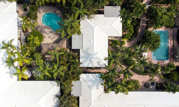 An aerial view of a resort with two swimming pools surrounded by palm trees and a white-roofed building.
