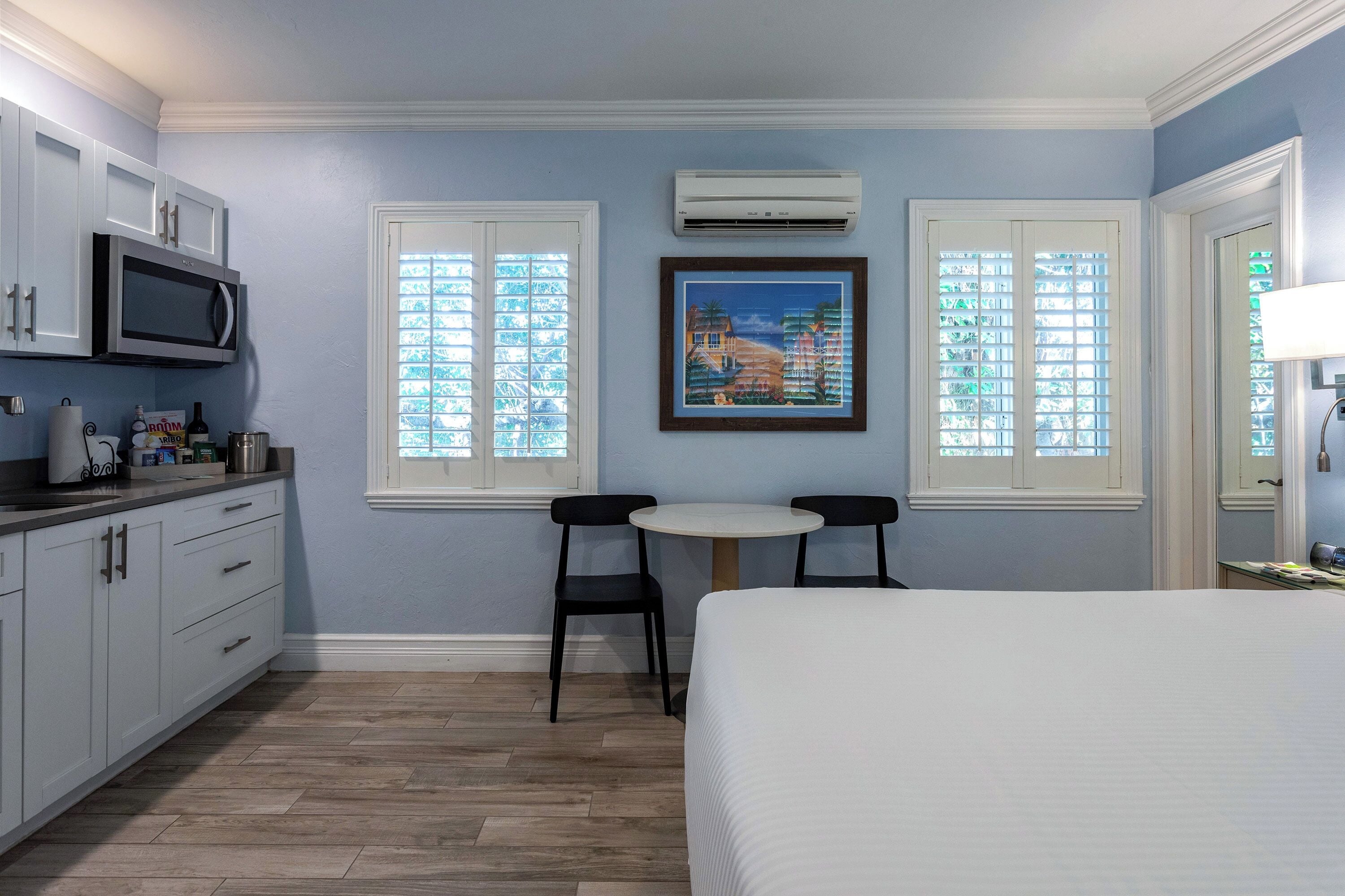 A bedroom with a bed, a desk, two chairs, a microwave, and a painting on the wall.