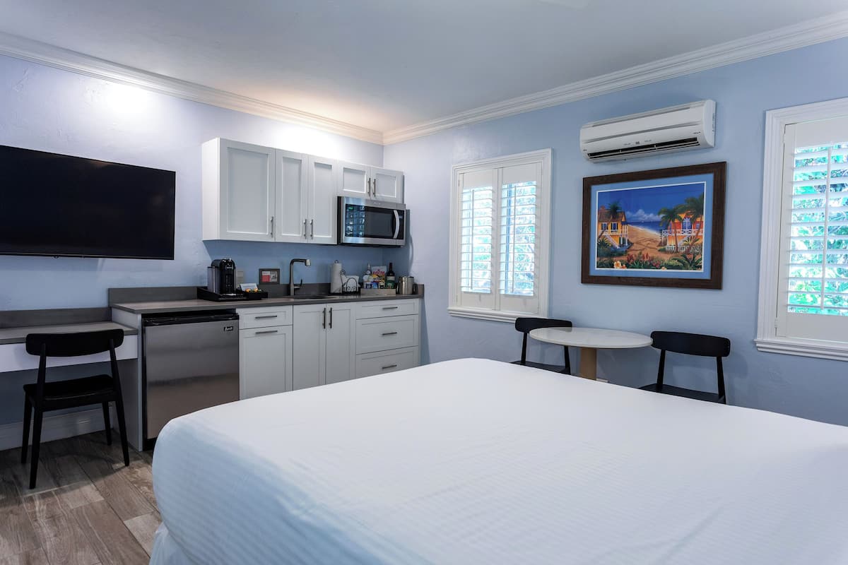 Ett hotellrum med en säng, ett skrivbord med en stol, ett litet kök, en tv och en tavla på väggen.