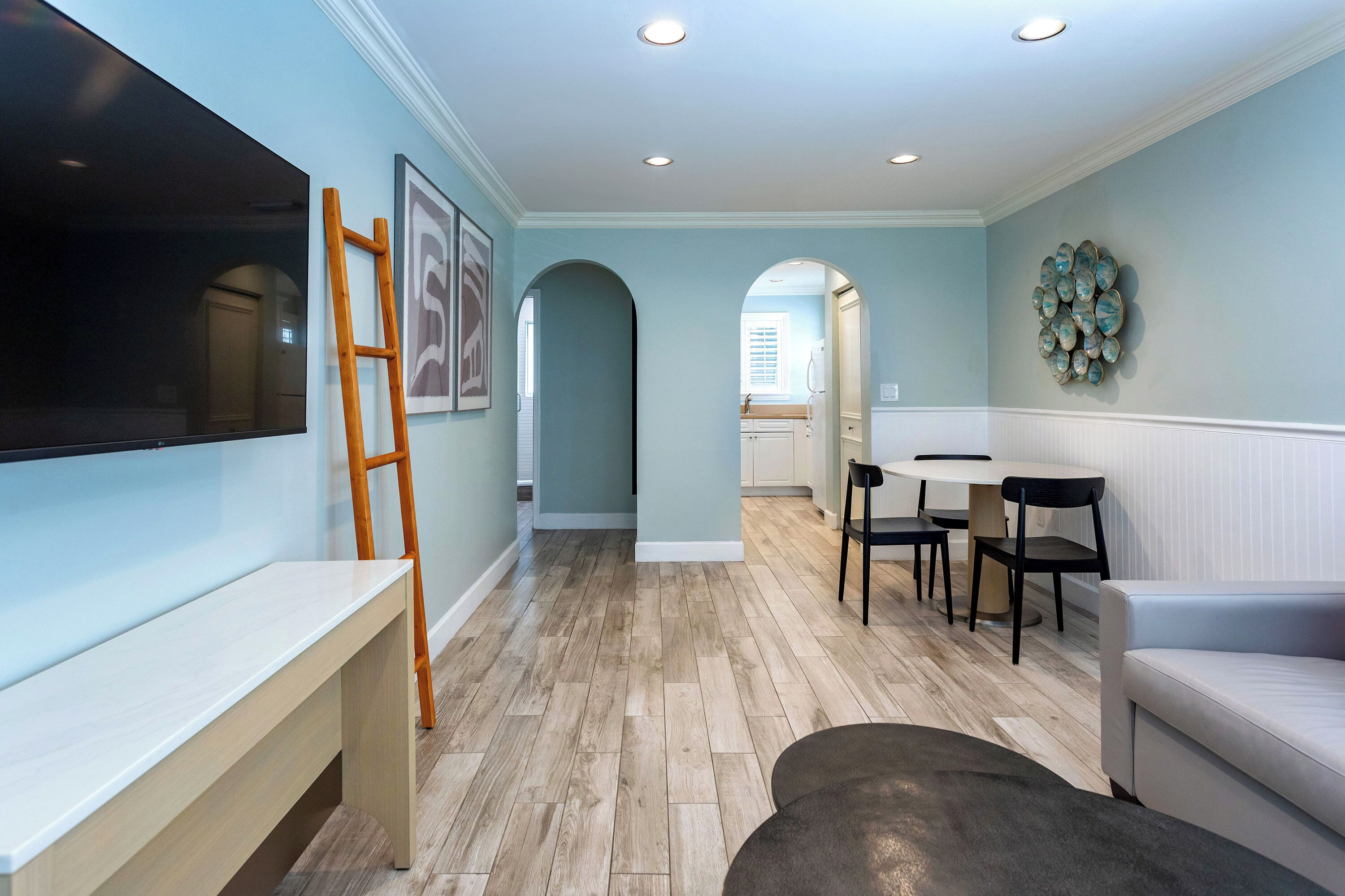 A modern living room with a flat-screen TV, a wooden ladder, a bench, a round table with chairs, and a light blue wall.