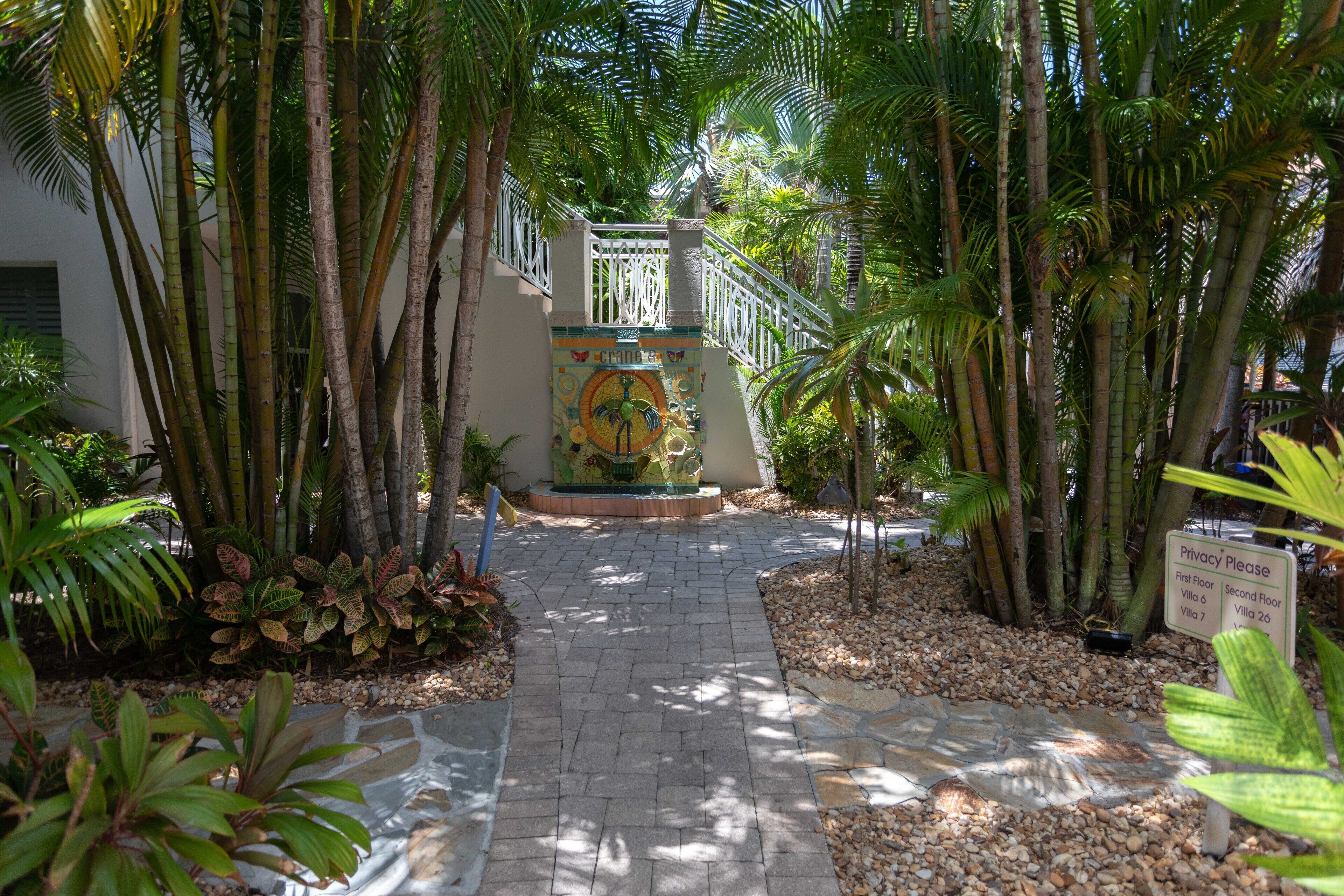 A paved pathway leading to a building with a mosaic mural, surrounded by palm trees and a privacy sign.
